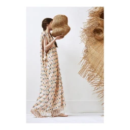 Woman in a patterned fringe dress holding a woven sculptural vessel against a white wall.