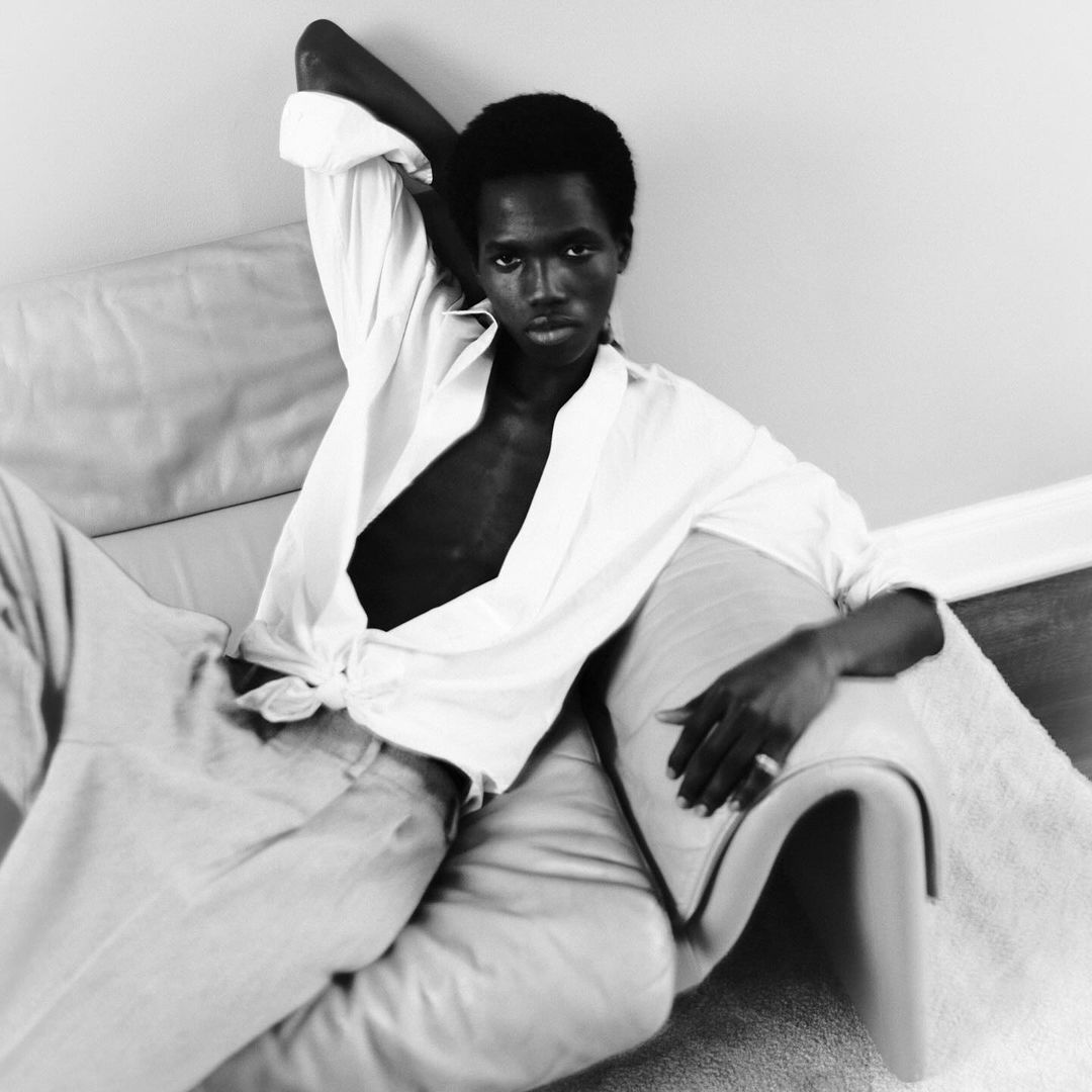 Monochrome portrait of a Black man reclining on a sofa in a white shirt and pants in a relaxed pose, gazing directly at the camera.
