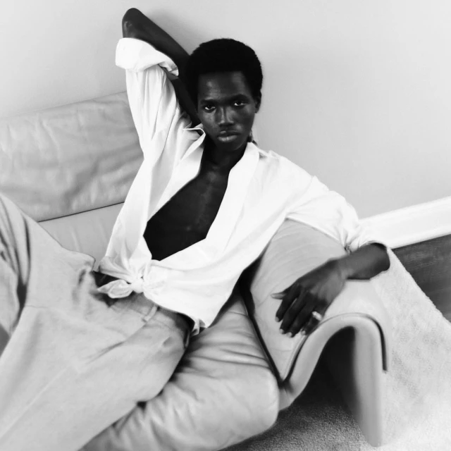 Monochrome portrait of a Black man reclining on a sofa in a white shirt and pants in a relaxed pose, gazing directly at the camera.