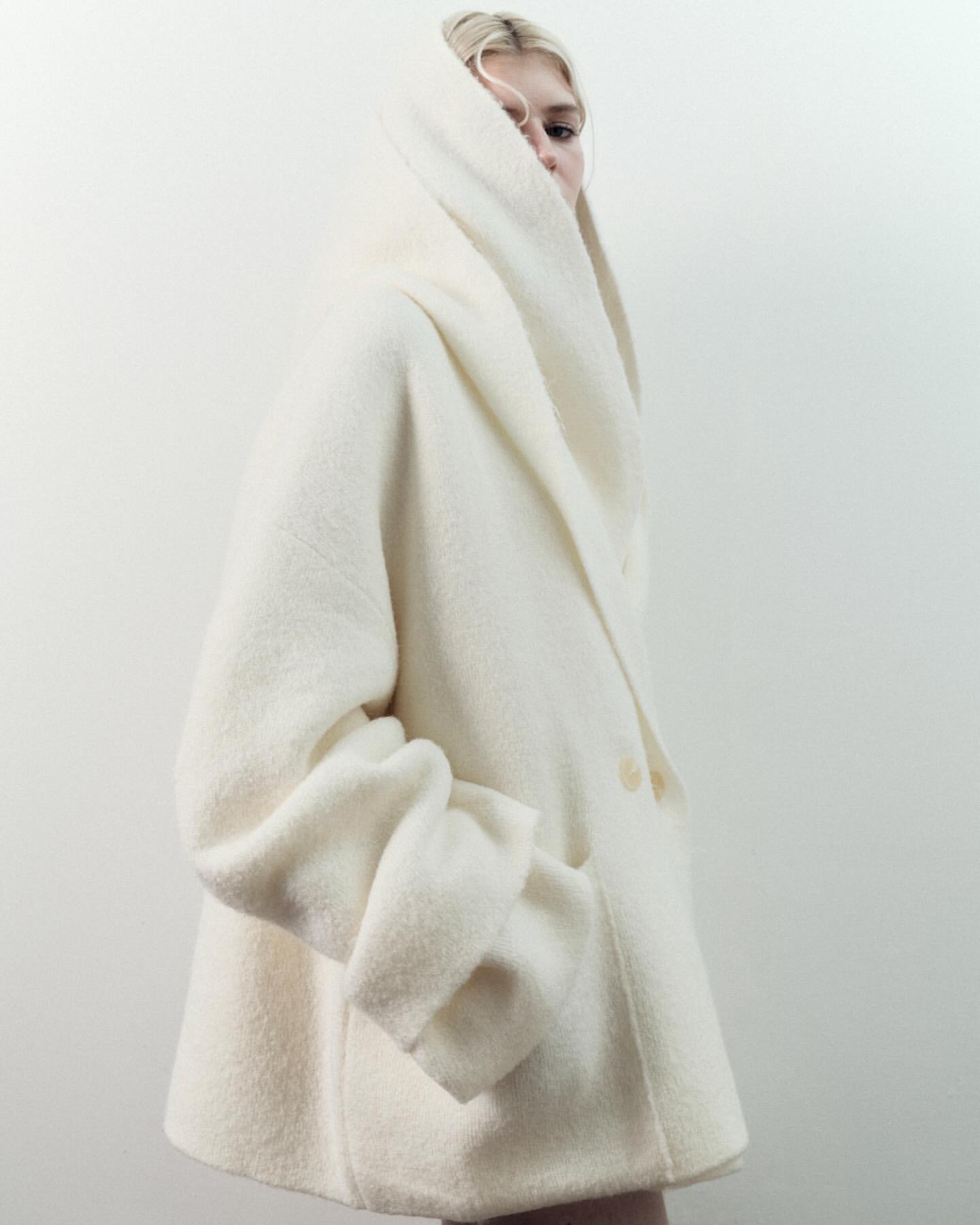 A blonde woman in a cream-colored, oversized wool coat with a hood poses against a white backdrop in a fashion-forward studio portrait.