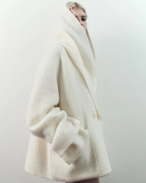 A blonde woman in a cream-colored, oversized wool coat with a hood poses against a white backdrop in a fashion-forward studio portrait.