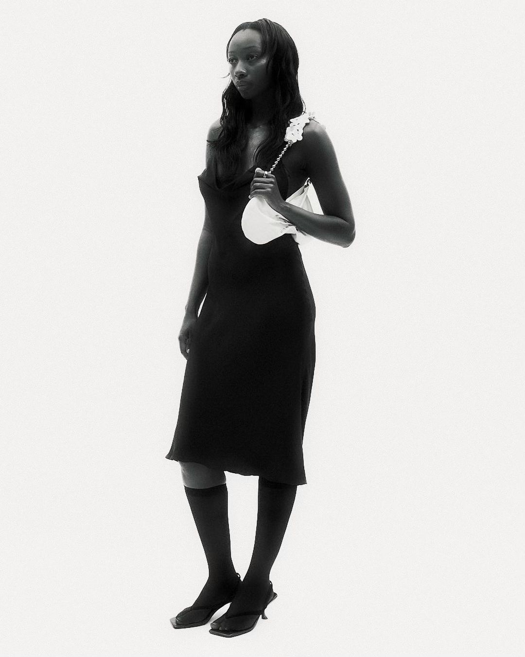Full-length studio shot of an adult woman posing in a black dress, knee-high socks, and heeled sandals, holding a white handbag.