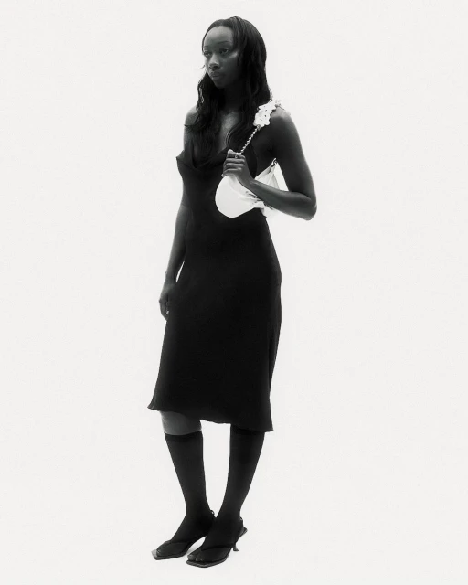 Full-length studio shot of an adult woman posing in a black dress, knee-high socks, and heeled sandals, holding a white handbag.