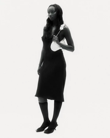 Full-length studio shot of an adult woman posing in a black dress, knee-high socks, and heeled sandals, holding a white handbag.