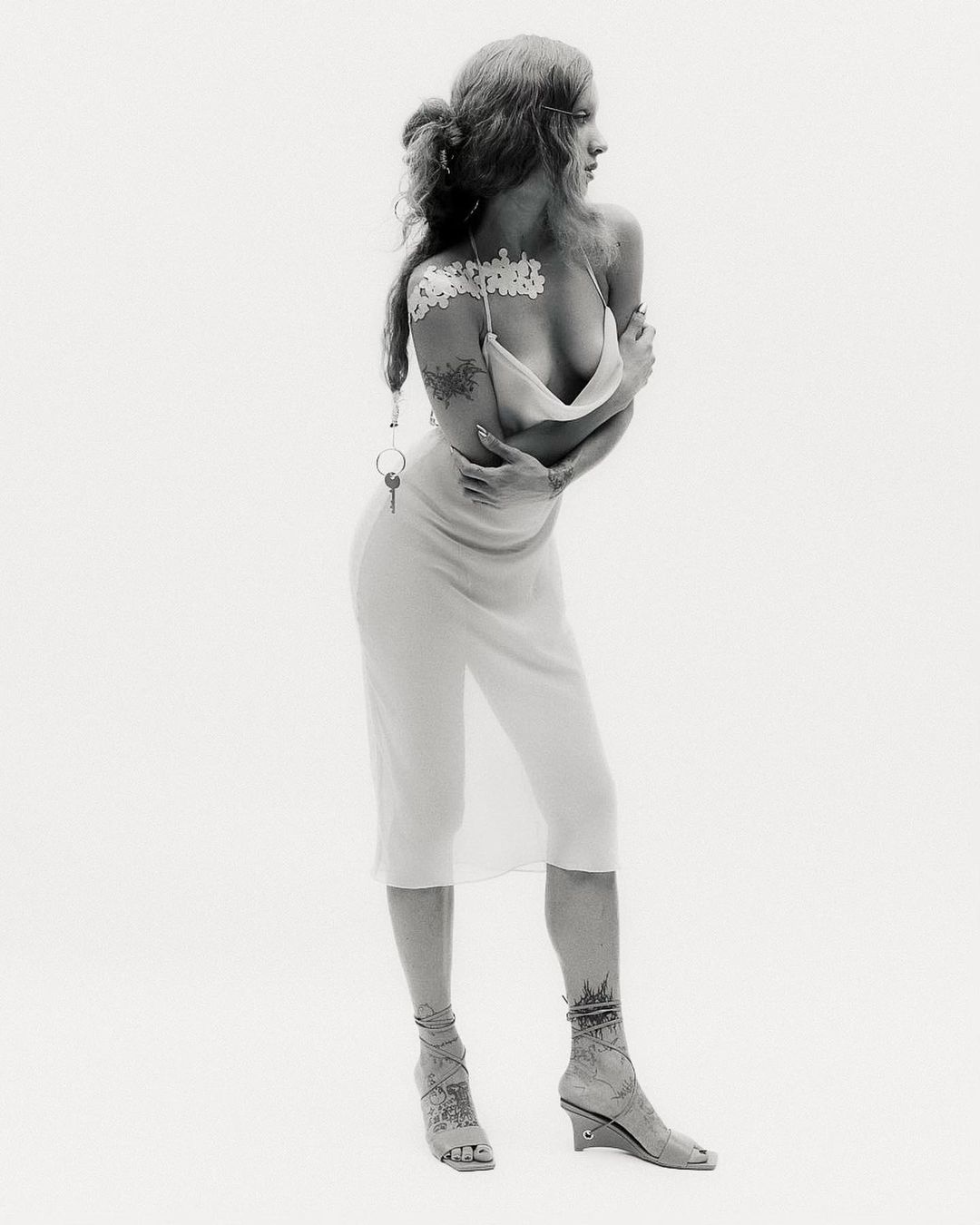 A full shot captures a slender adult female model in a semi-nude fashion pose, arms crossed, against a plain white backdrop.