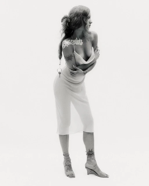 A full shot captures a slender adult female model in a semi-nude fashion pose, arms crossed, against a plain white backdrop.
