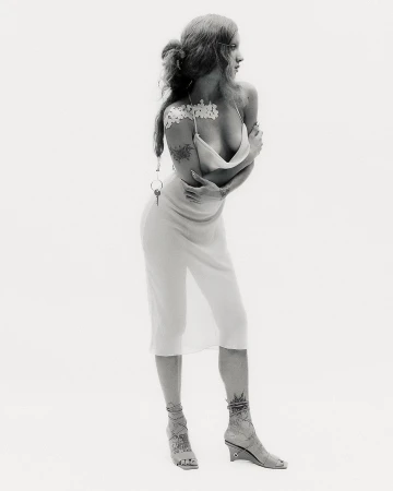 A full shot captures a slender adult female model in a semi-nude fashion pose, arms crossed, against a plain white backdrop.