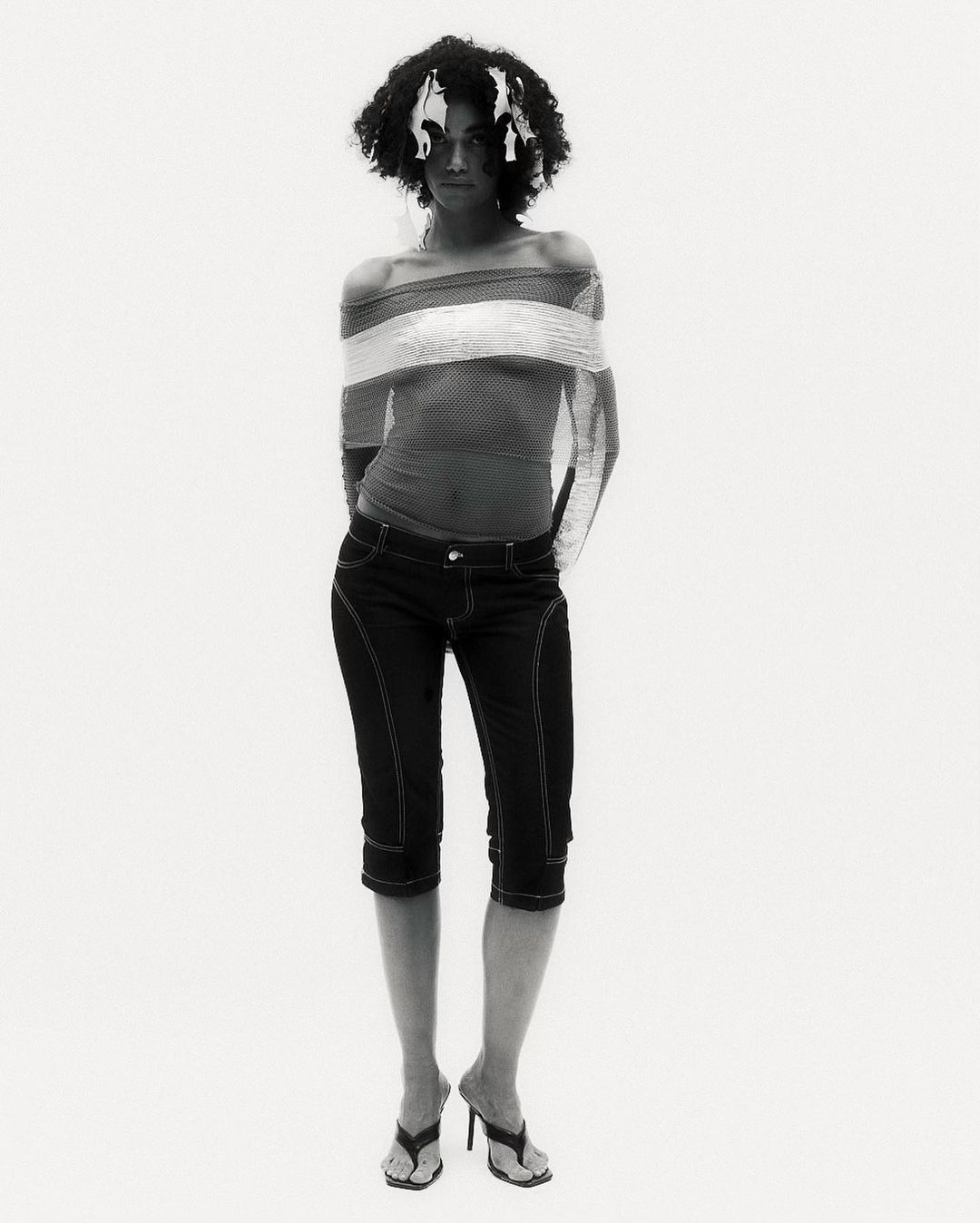 Woman with afro posing in off-the-shoulder top, cropped jeans, and heeled sandals in a full shot against a white studio backdrop.