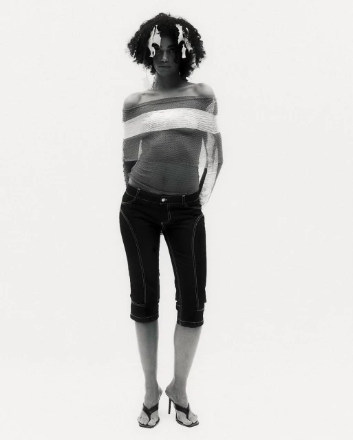 Woman with afro posing in off-the-shoulder top, cropped jeans, and heeled sandals in a full shot against a white studio backdrop.