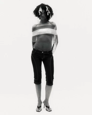 Woman with afro posing in off-the-shoulder top, cropped jeans, and heeled sandals in a full shot against a white studio backdrop.