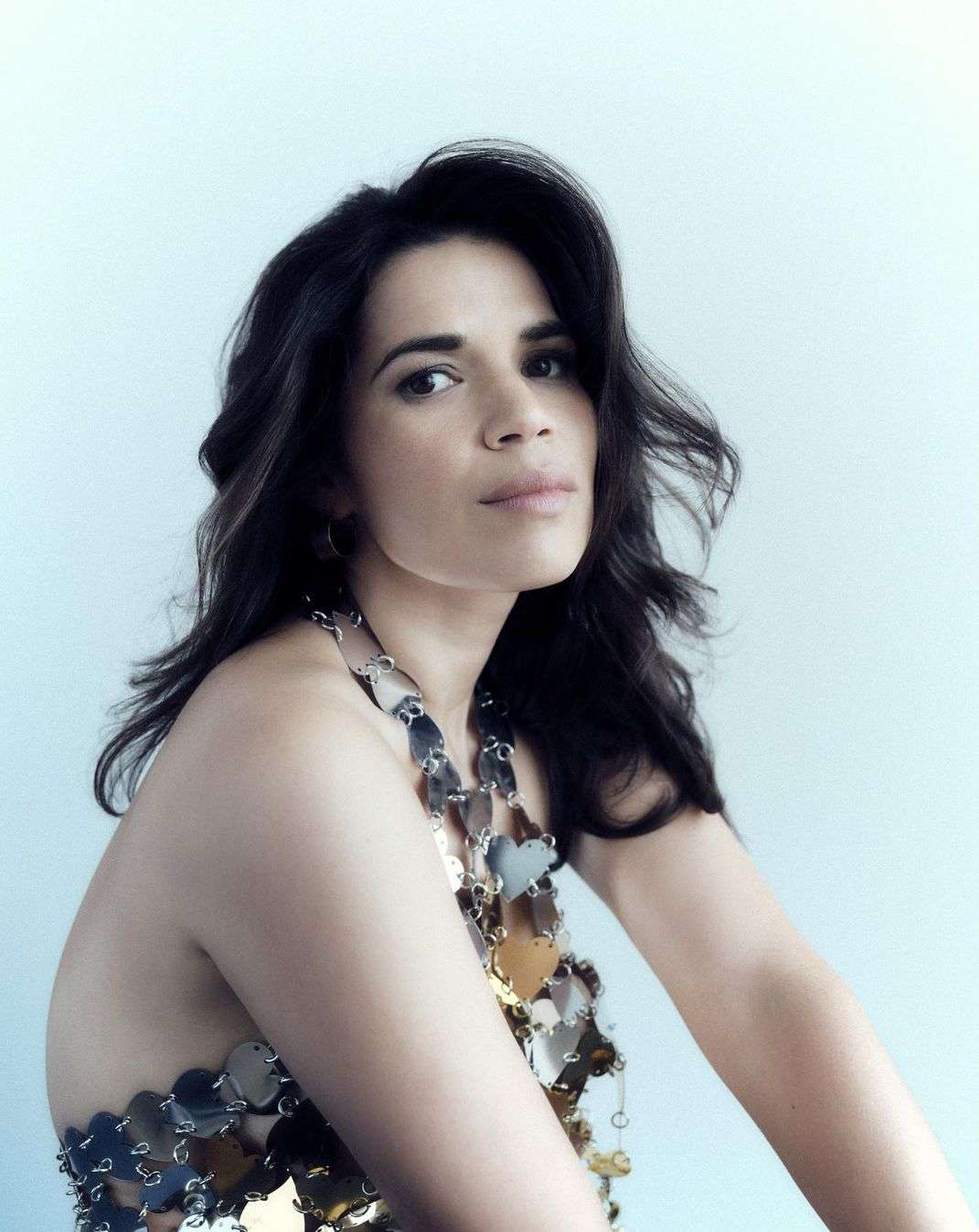 Portrait of a woman with dark curly hair wearing a silver, heart-shaped halter top against a light blue gradient background.