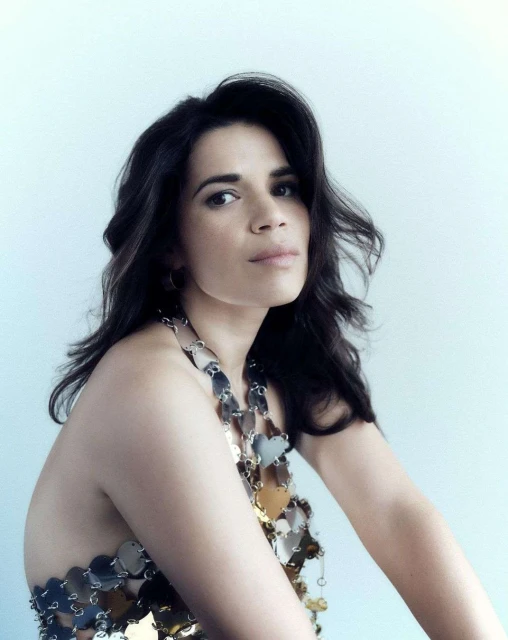 Portrait of a woman with dark curly hair wearing a silver, heart-shaped halter top against a light blue gradient background.