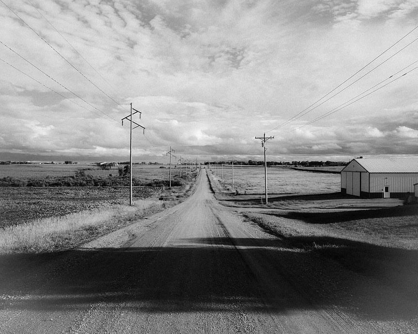 Monochrome landscape featuring a long dirt road leading to the horizon, flanked by fields, utility poles, and a barn under a cloudy sky.