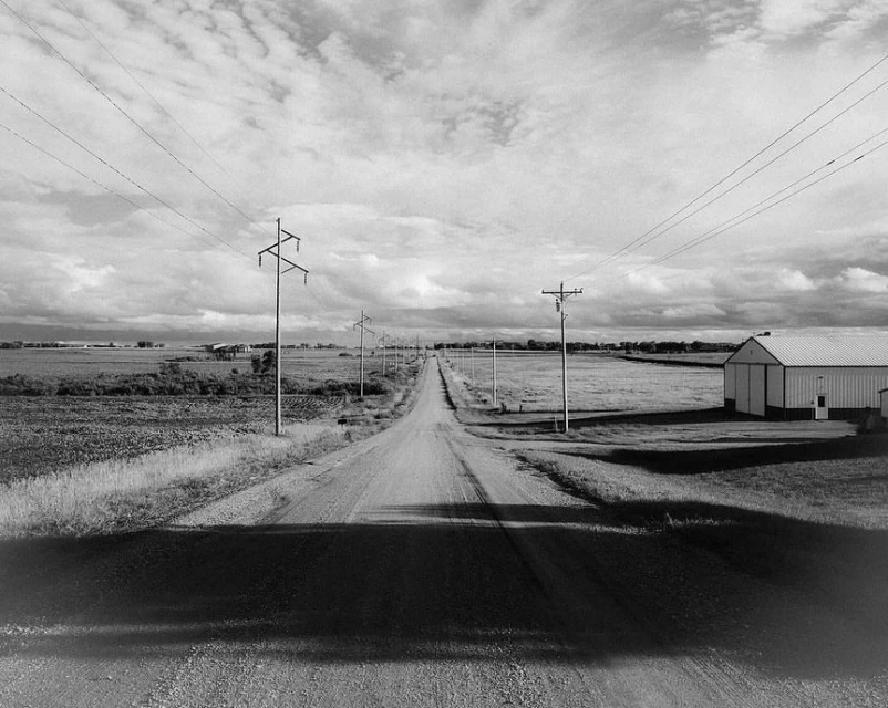 Monochrome landscape featuring a long dirt road leading to the horizon, flanked by fields, utility poles, and a barn under a cloudy sky.