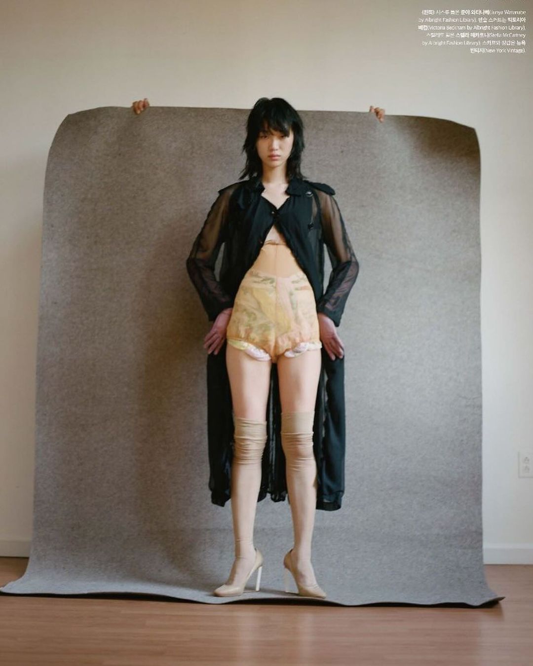 A woman stands in a photo studio wearing a black coat, beige shorts and matching thigh-high stockings. The background is beige.