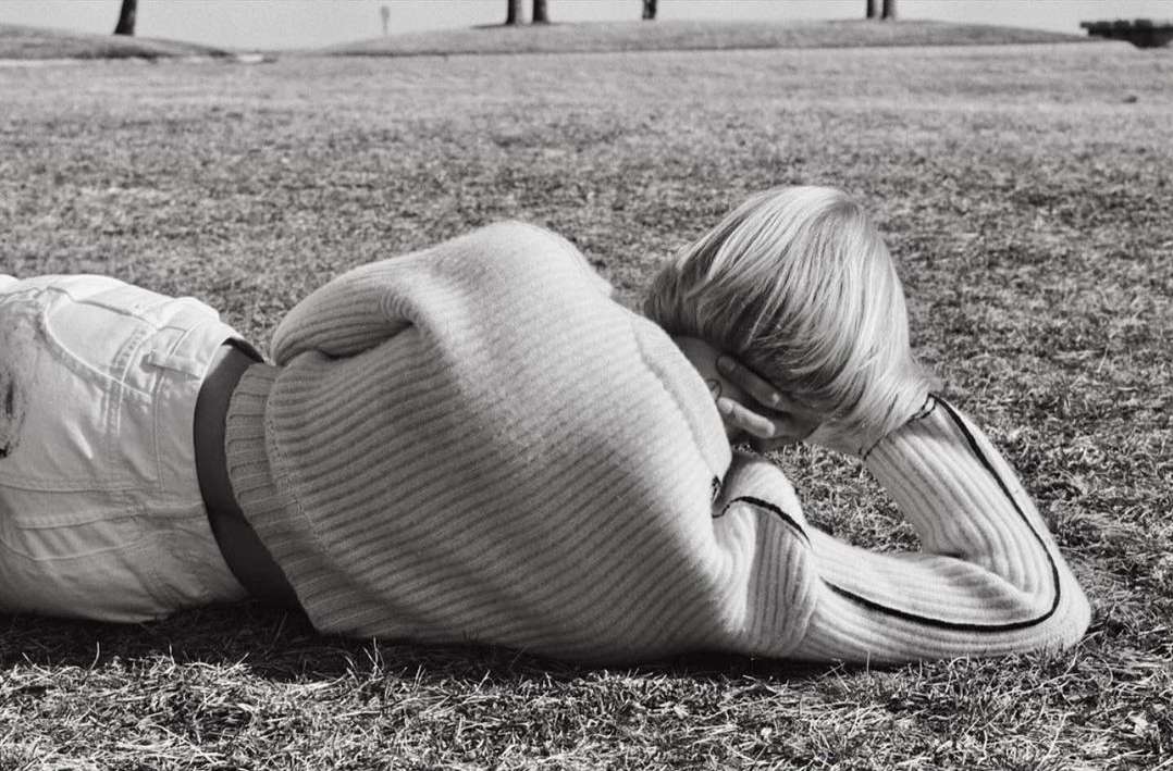A blonde person wearing a ribbed sweater lying prone on grassy ground under natural daylight