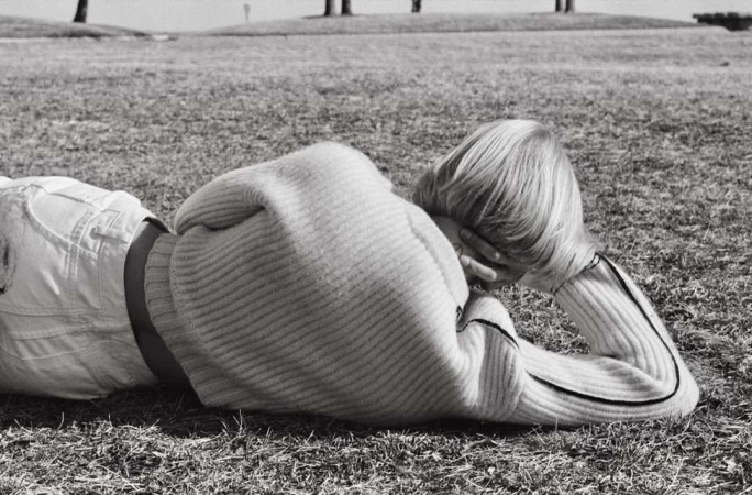 A blonde person wearing a ribbed sweater lying prone on grassy ground under natural daylight