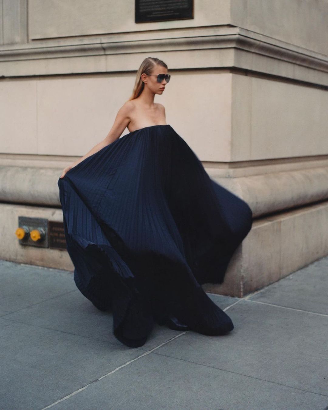 A fashion shot shows a woman with blonde hair wearing sunglasses and a large, pleated navy dress, walking on a city sidewalk.