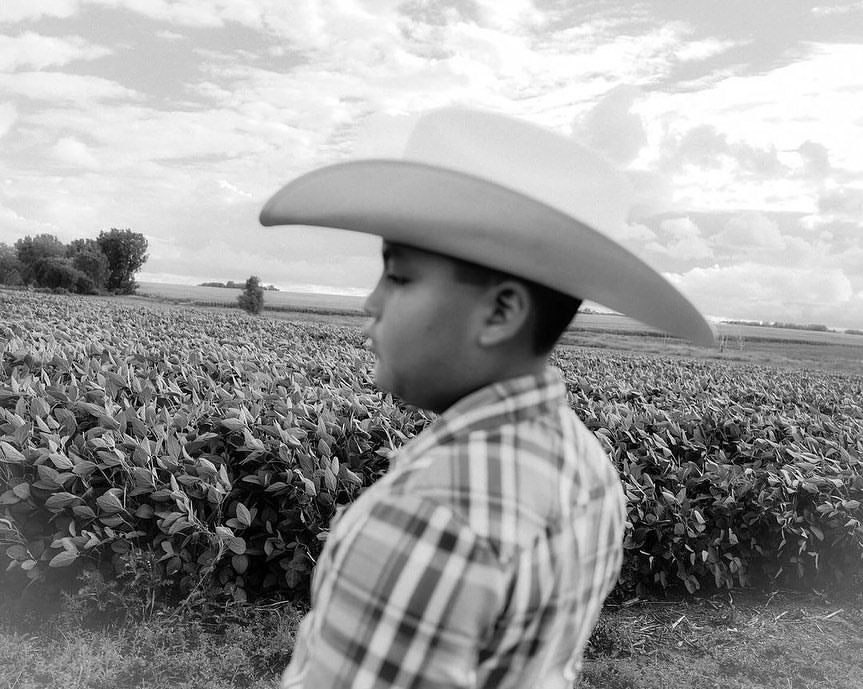 Black and white portrait of a young person in a cowboy hat and plaid shirt, standing in a field under a cloudy sky, side view.