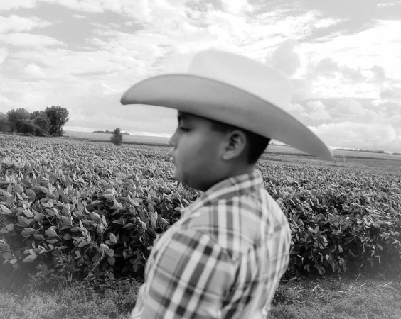 Black and white portrait of a young person in a cowboy hat and plaid shirt, standing in a field under a cloudy sky, side view.