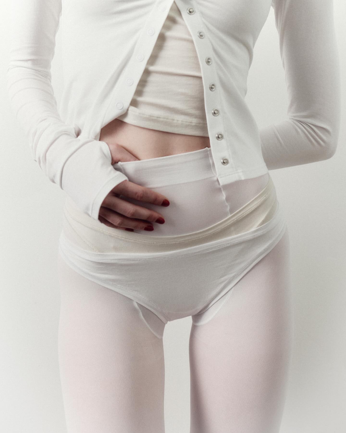 Cropped shot of a fair-skinned young adult woman in white top and bottom, holding stomach in an indoor studio setting.