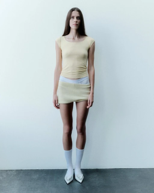 Full shot of a young adult woman posing in a light yellow shirt and skirt, white socks and shoes, against a white wall backdrop.