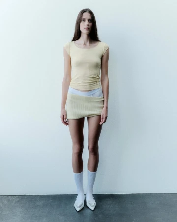 Full-length portrait of a female model wearing a pale yellow top and miniskirt with white socks and pointed heels against a white studio wall.