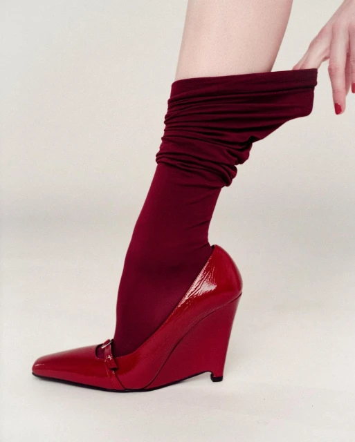 A woman's leg is adorned with a red sock and a shiny red wedge heel shoe, posed against a clean, bright white background.