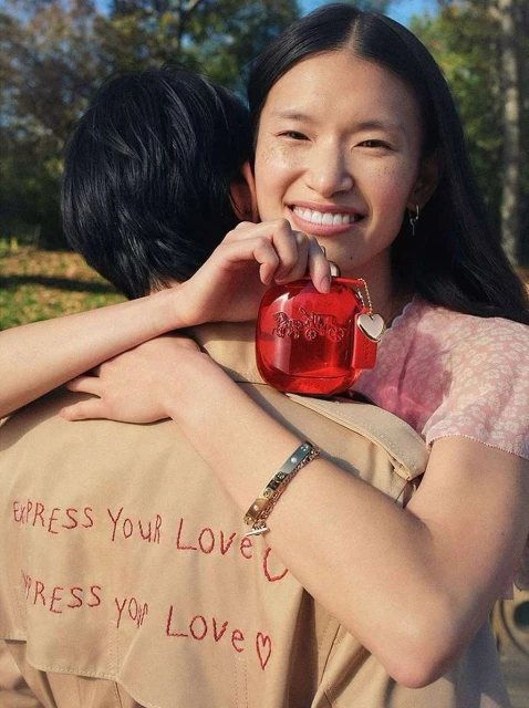 A smiling woman holds red perfume bottle while being embraced by another person outdoors in a sunlit park setting with green trees.
