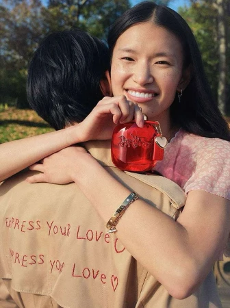 A smiling woman holds red perfume bottle while being embraced by another person outdoors in a sunlit park setting with green trees.