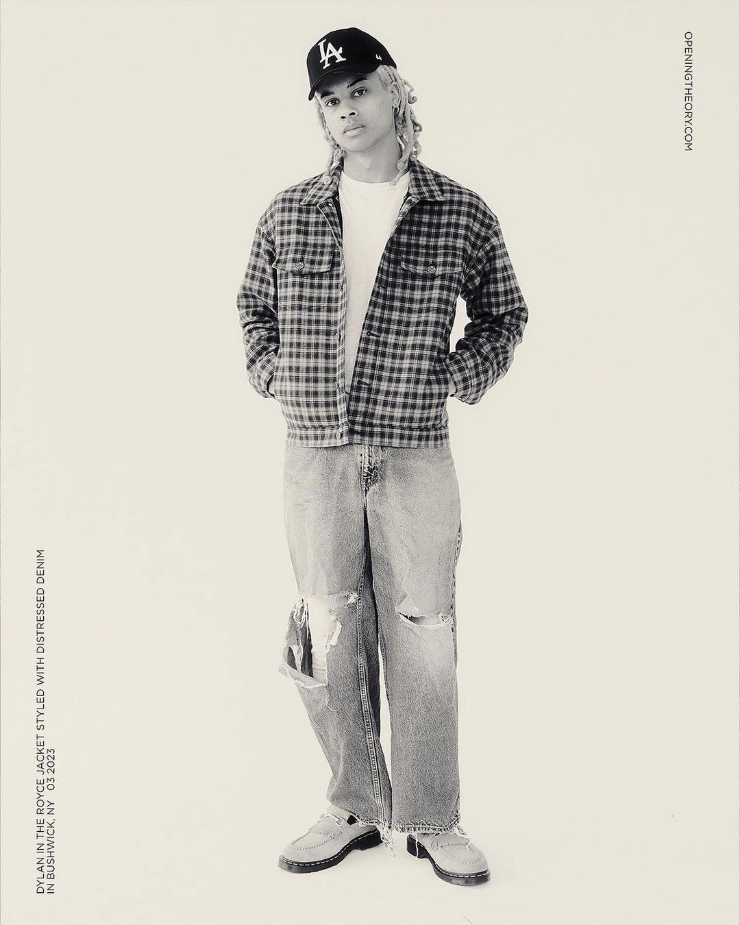 Full-length studio shot of a young adult wearing a baseball cap, jacket, t-shirt, distressed denim jeans, and moccasins against a neutral backdrop.