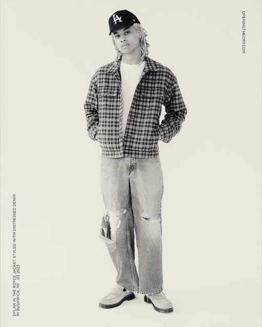 Full-length studio shot of a young adult wearing a baseball cap, jacket, t-shirt, distressed denim jeans, and moccasins against a neutral backdrop.