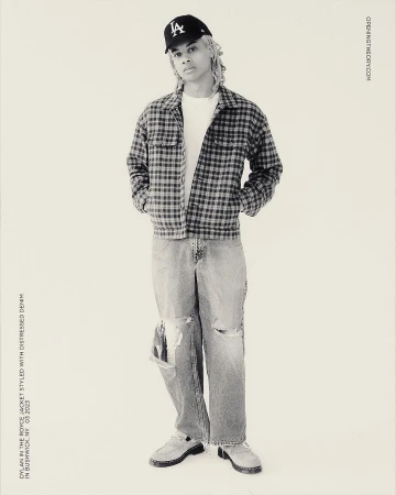 Full-length studio shot of a young adult wearing a baseball cap, jacket, t-shirt, distressed denim jeans, and moccasins against a neutral backdrop.