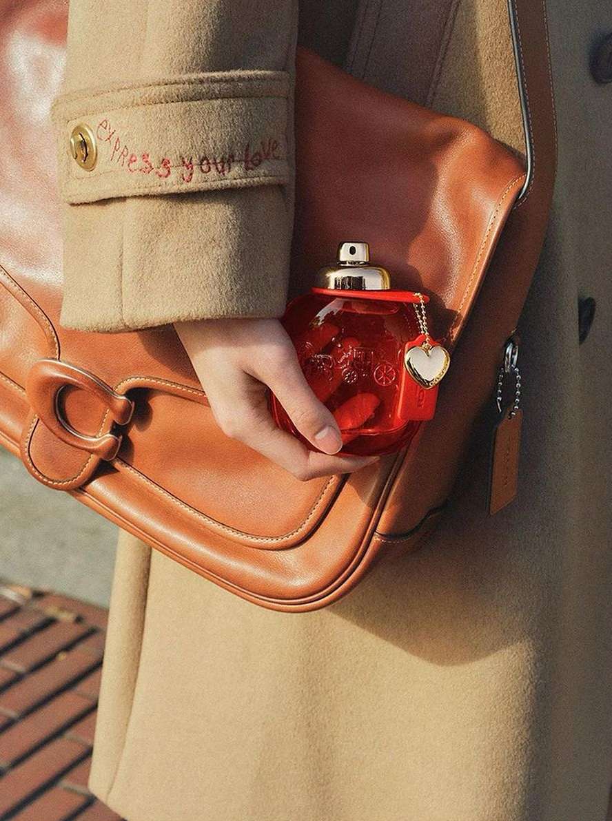 A woman holds a red perfume bottle shaped like a purse in front of a Coach handbag and a tan overcoat in a soft, natural light.