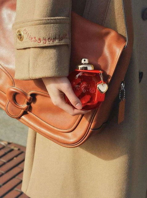 A woman holds a red perfume bottle shaped like a purse in front of a Coach handbag and a tan overcoat in a soft, natural light.