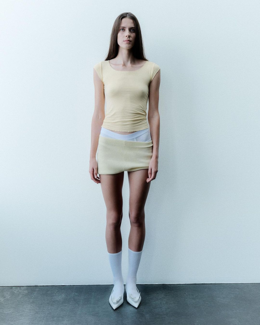 Full-length studio shot of a woman in a yellow top and skirt, wearing white socks and heels, posing against a plain off-white wall.