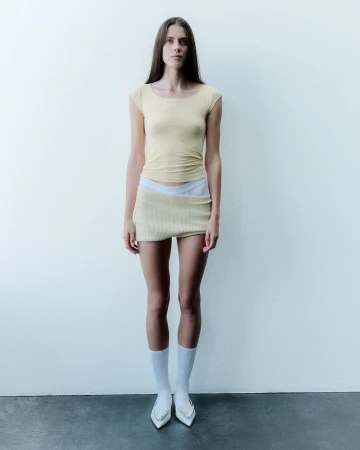 Full-length studio shot of a woman in a yellow top and skirt, wearing white socks and heels, posing against a plain off-white wall.
