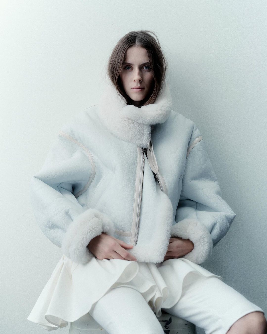 A woman with long brown hair sits in a studio wearing a light blue bomber jacket with a white fur collar and cuffs and white pants.