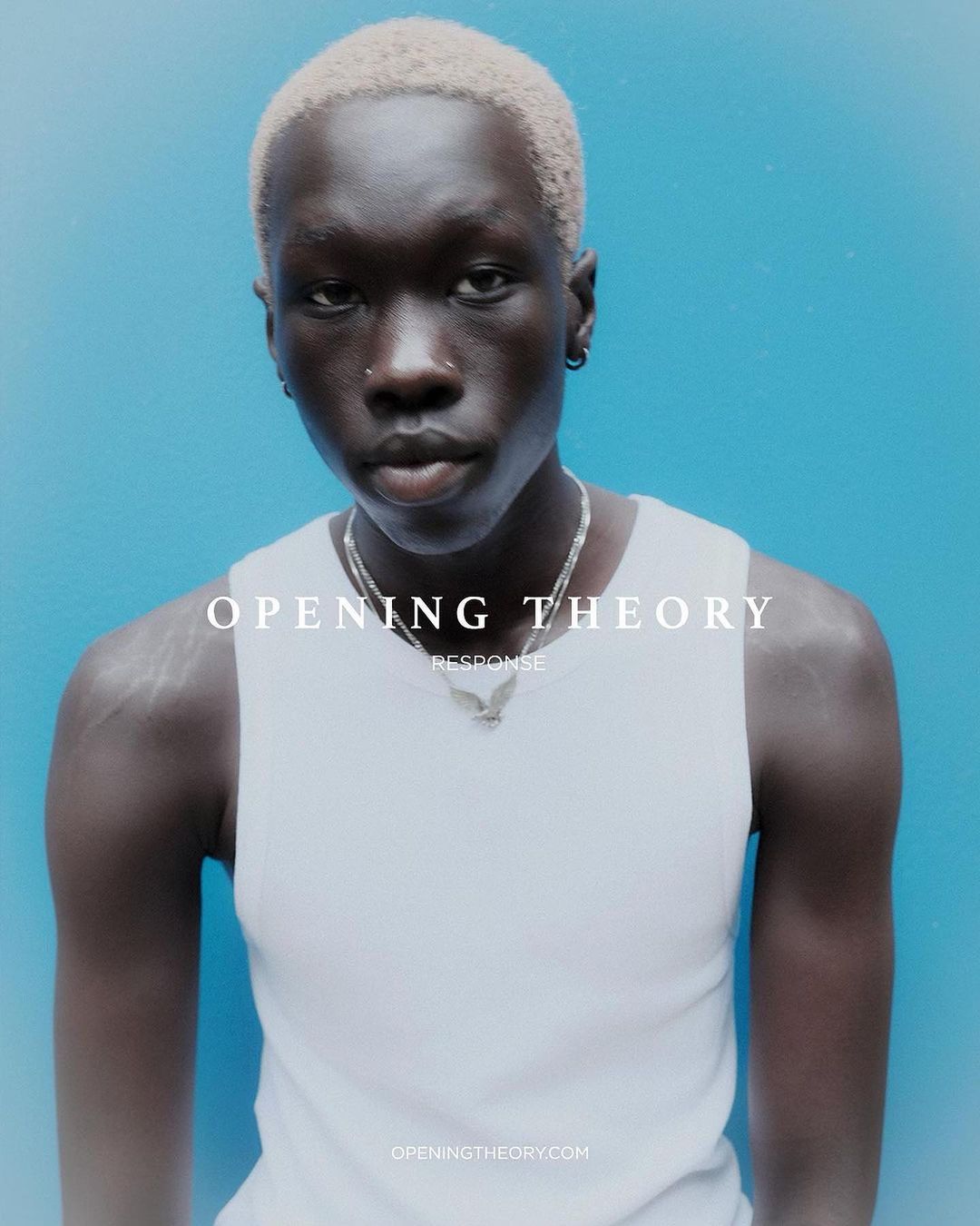 Portrait of a young adult with bleached blonde hair and silver chain against a bright blue background in a minimalist fashion shot.