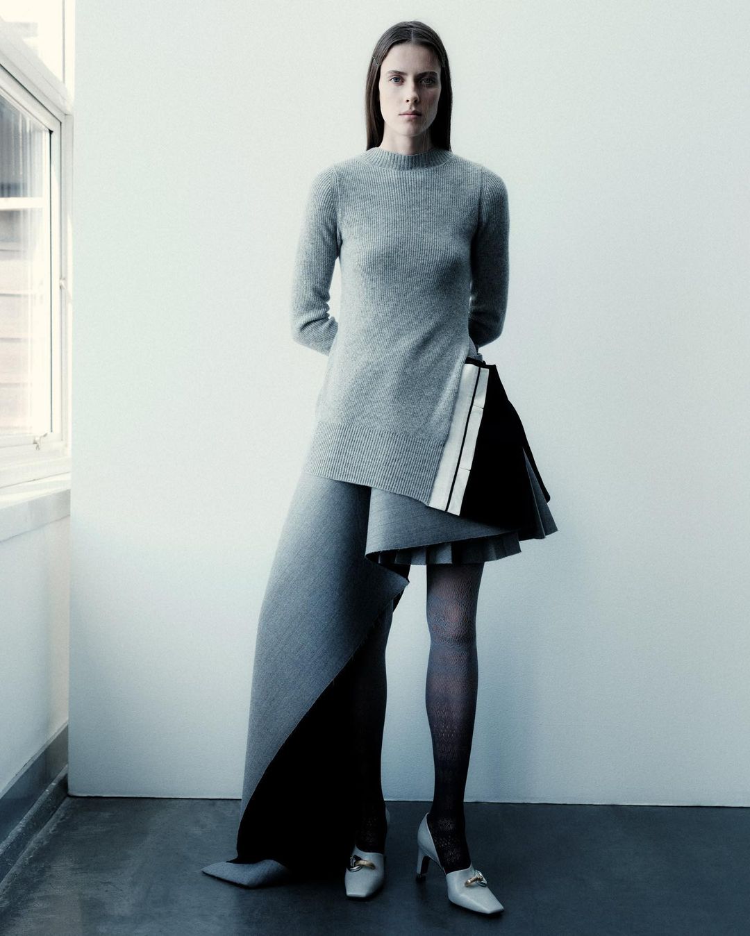 Fashion shot of a slender woman in a grey sweater dress with black and white accents, standing against a white wall near a window.