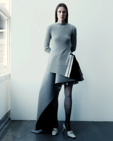 Fashion shot of a slender woman in a grey sweater dress with black and white accents, standing against a white wall near a window.