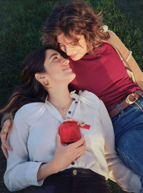 Two women lie on grass, smiling at each other. One wears a white blouse and holds a red perfume bottle; the other wears a maroon shirt.