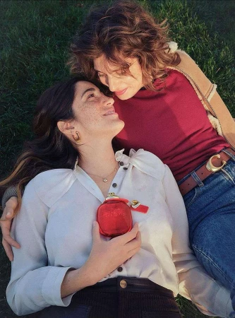 Two women lie on grass, smiling at each other. One wears a white blouse and holds a red perfume bottle; the other wears a maroon shirt.