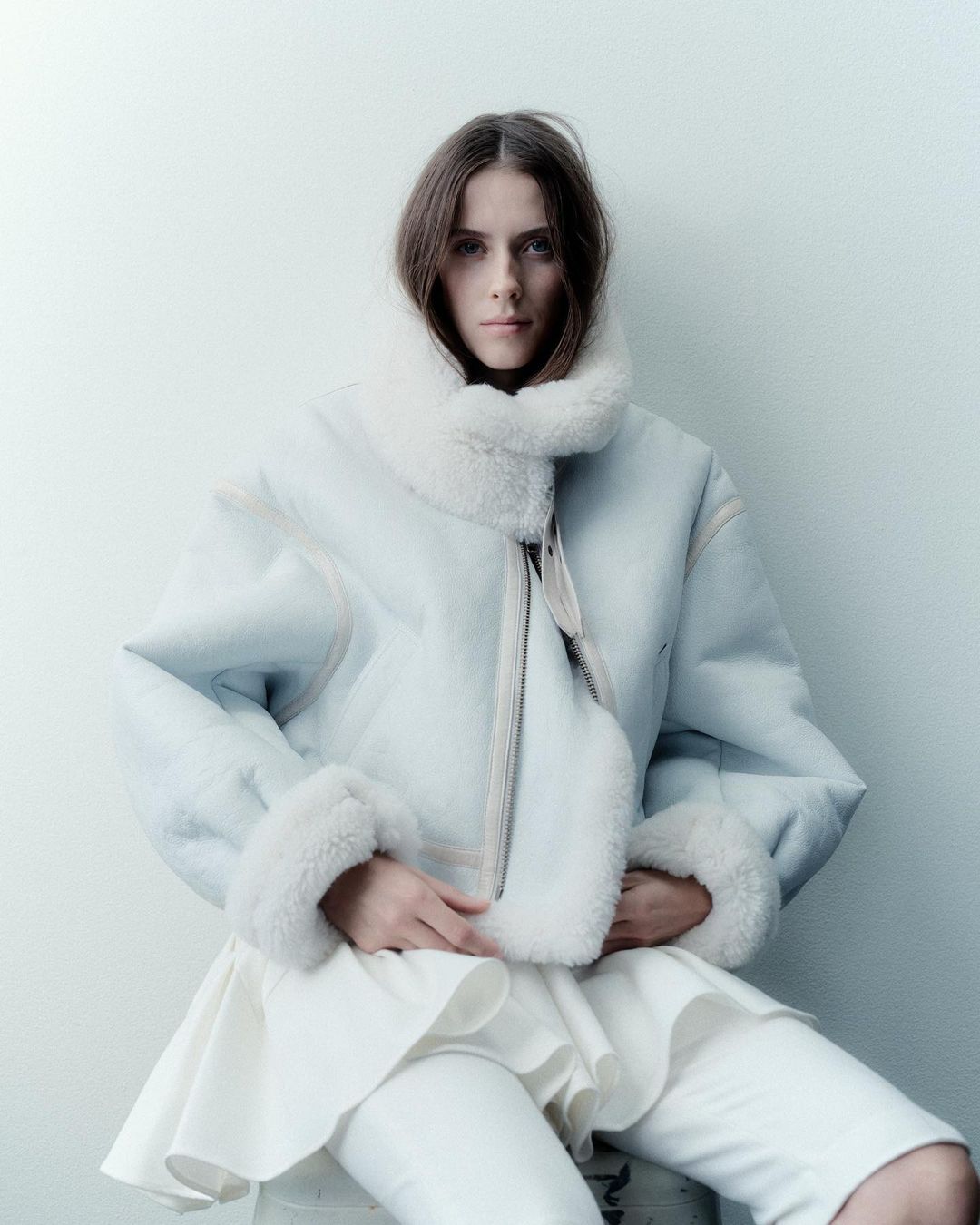 A young woman with dark blonde hair poses indoors wearing an oversized, pale blue shearling jacket over a layered white outfit.