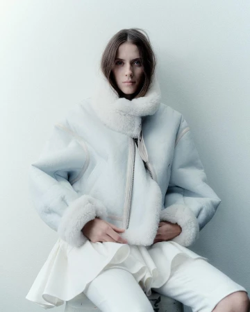 A young woman with dark blonde hair poses indoors wearing an oversized, pale blue shearling jacket over a layered white outfit.
