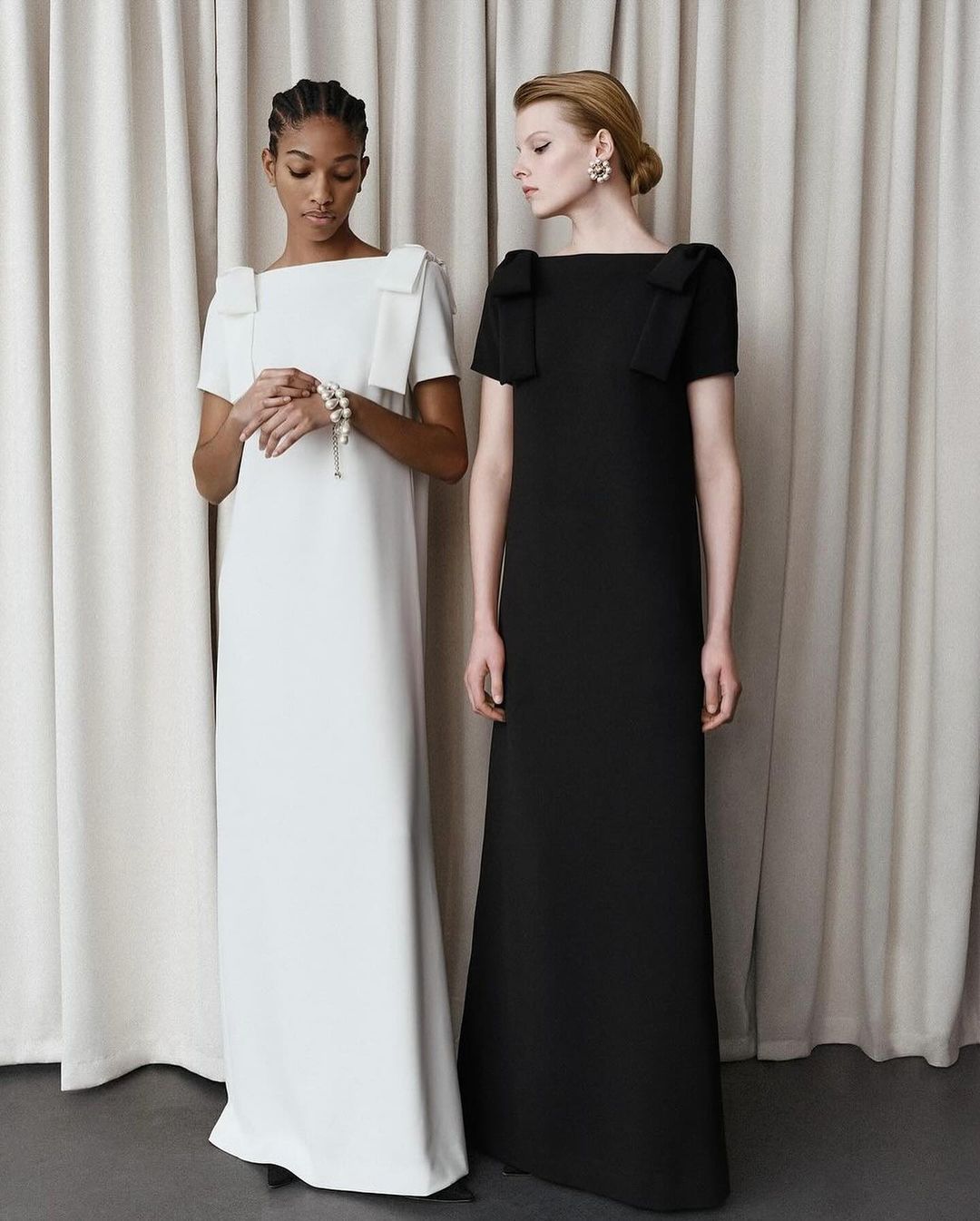 Two women stand in a photo studio, modeling elegant floor-length evening gowns with square necklines and bow details, against a cream backdrop.