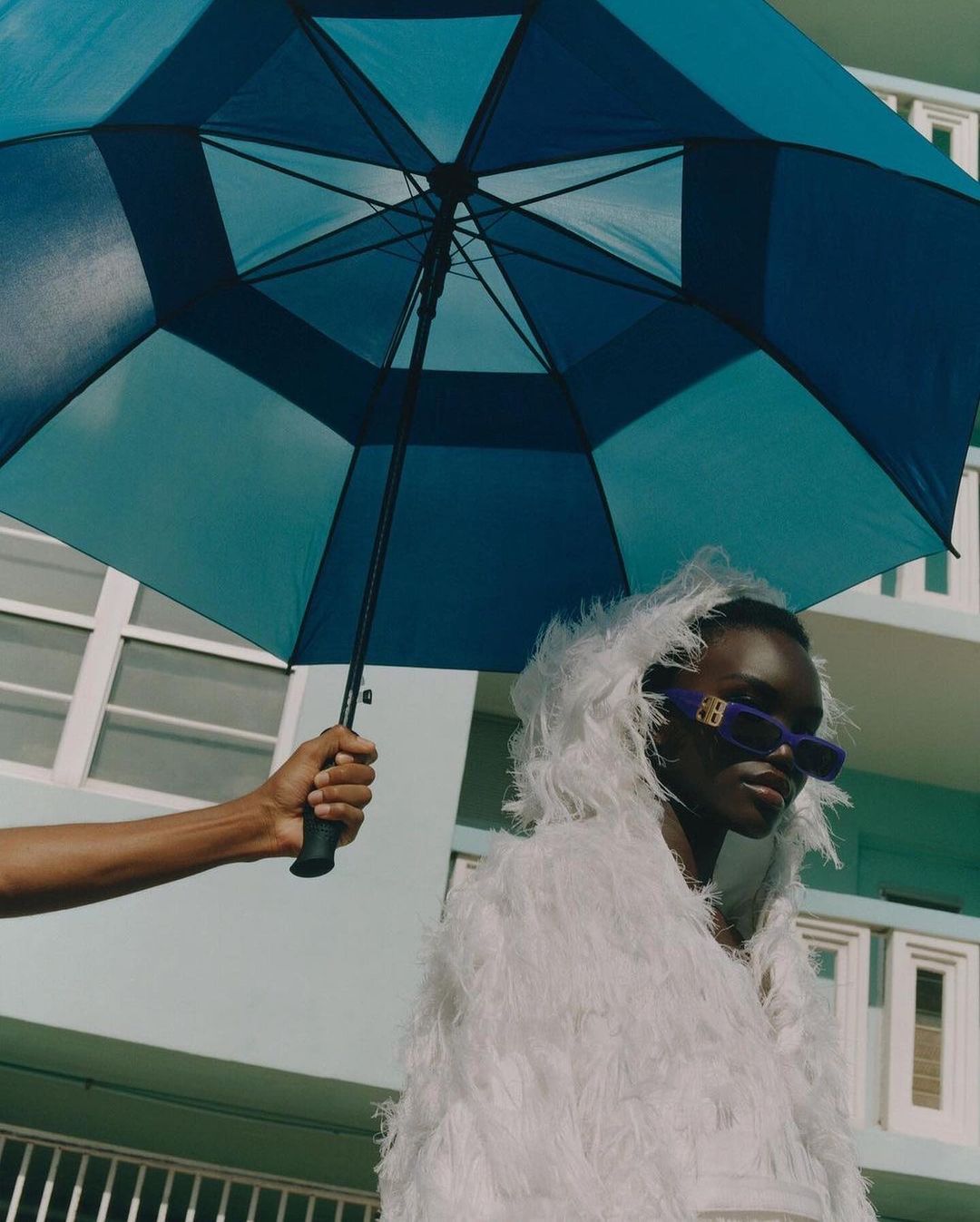 Stylish portrait of an adult female under a blue umbrella outdoors, wearing purple sunglasses and a white feathery jacket.