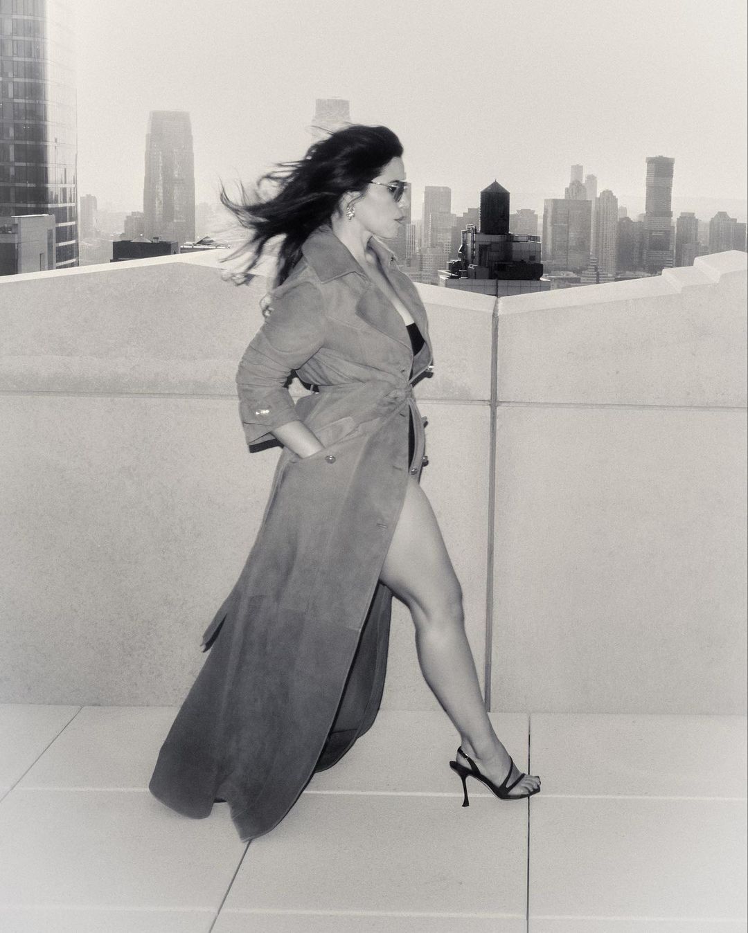 A woman in a trench coat and heels walks on a rooftop with a city skyline in the background, captured in black and white.