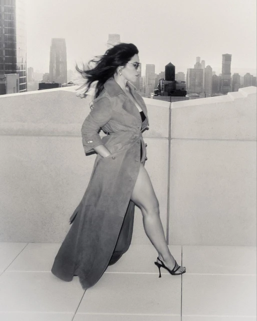 A woman in a trench coat and heels walks on a rooftop with a city skyline in the background, captured in black and white.