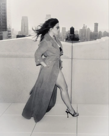A woman in a trench coat and heels walks on a rooftop with a city skyline in the background, captured in black and white.